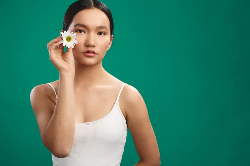 asian woman holds a white flower near her face in a clean studio scene, calm atmosphere, soft lighting, and a bold green backdrop that enhances simplicity and focus on natural expression.