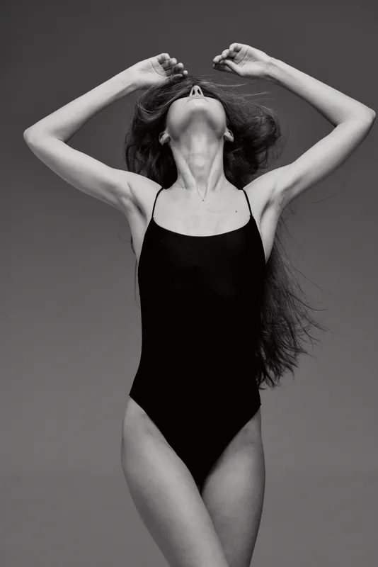 Black and white elegant woman in swimsuit stretching with raised arms against plain background.