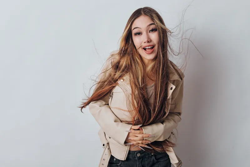 Young woman with long hair and beige jacket posing against gray background with playful expression. Casual style, natural makeup, and confident attitude for lifestyle or fashion concept.