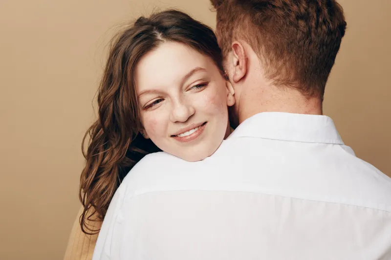 Smiling woman with long hair embraces man in white shirt, showing affection and warmth. Close-up on beige background. Concept of love, relationship, and intimacy between couple.