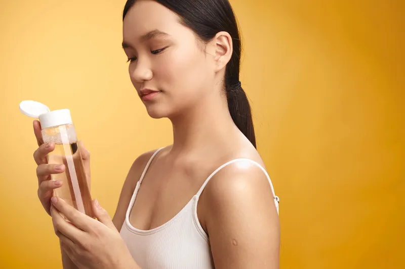 asian woman examines a cosmetic bottle in a bright studio, focusing on skincare product details while gentle light highlights her calm, confident expression.
