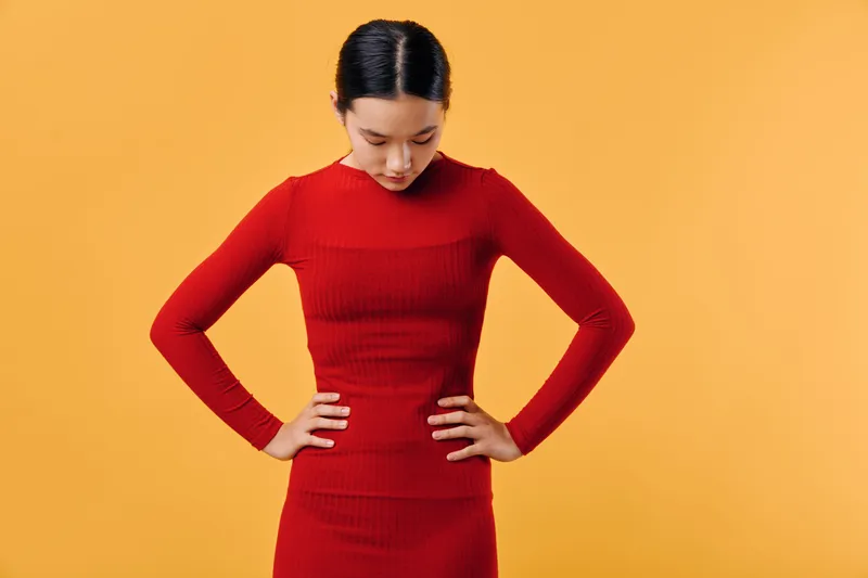 Woman in red dress looking down with hands on hips standing against orange background. Fashion, confidence, pose, female model with casual style and minimalistic studio portrait concept.