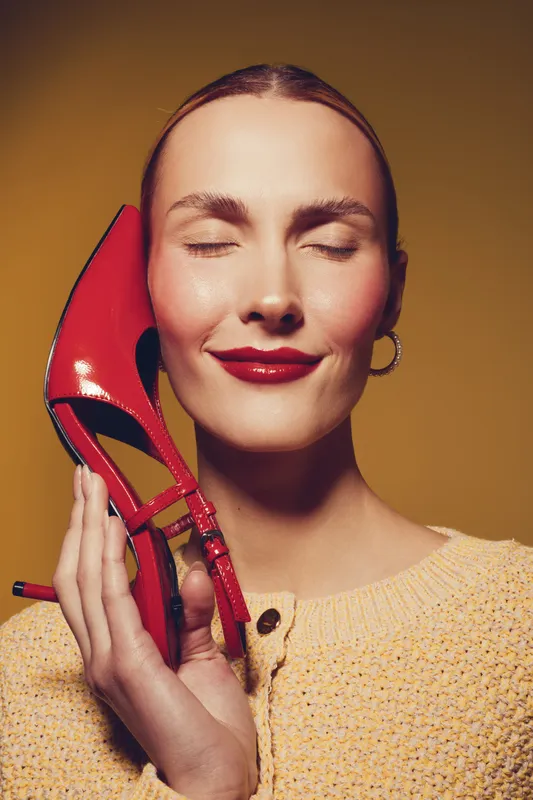 Smiling woman with closed eyes holds red high heel shoe near face. Warm yellow background and soft lighting create cozy mood. Concept of fashion, beauty, and style accessory.