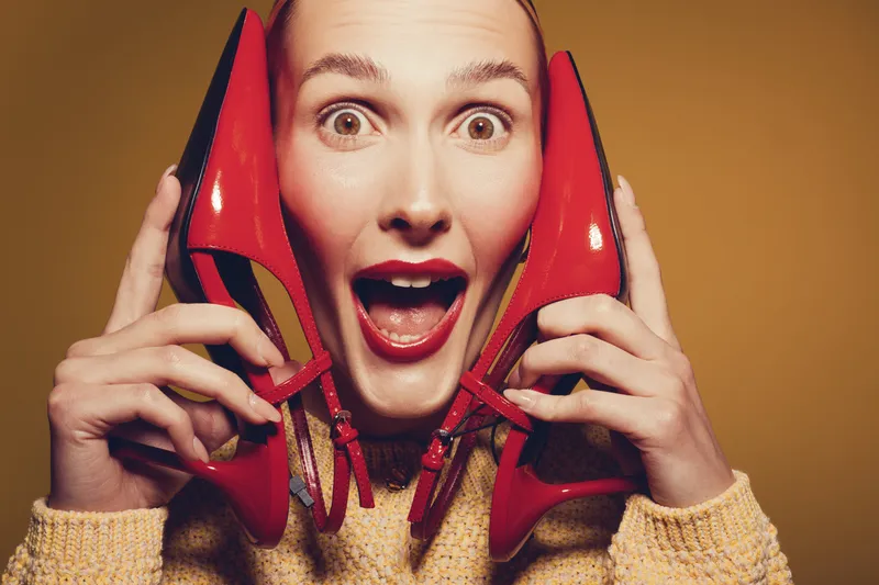 Excited woman holds red high heels near face with wide eyes and open mouth. Studio portrait on brown background. Concept for fashion, shoes, and joyful emotions.