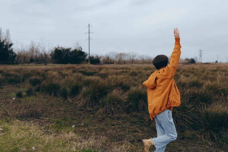 child in orange raincoat outdoors in grassy field, hands raised while jumping, playful lifestyle moment in spring air, windy overcast sky, nature landscape, carefree child freedom now