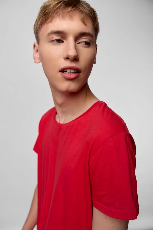 Young man with a red shirt turns slightly to the side, showcasing casual fashion and modern styling in a simple studio setting, with natural lighting and a relaxed mood