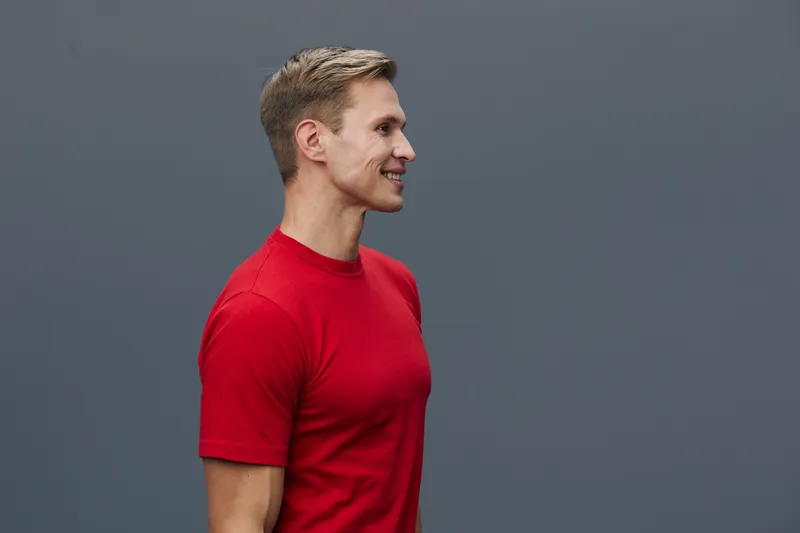 Smiling man wearing red t-shirt standing sideways against a plain gray background in a professional studio photo. Happy, confident, casual, portrait, fashion, lifestyle, positive energy.