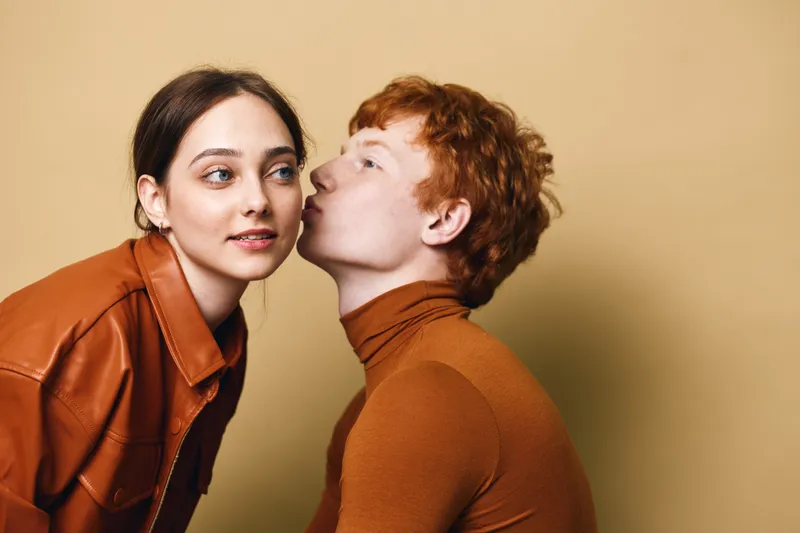 Young man kissing woman on cheek with beige background. Woman in brown leather jacket looking away with smile. Romance, affection, closeup, casual fashion moment.