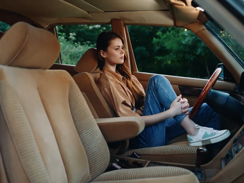 Young woman in casual outfit reflecting in vintage car interior, evoking feelings of nostalgia and tranquility, set against a lush green backdrop.