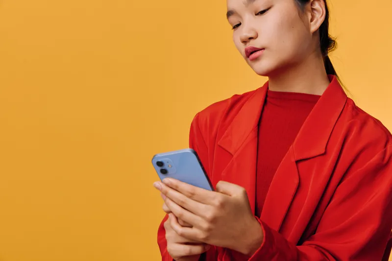 Woman in red jacket holding smartphone and looking at screen with focused expression, isolated on yellow background. Technology, communication, mobile and connection concept.