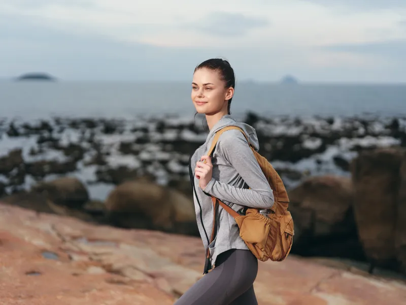 Young woman enjoying a hike by the seaside, wearing casual activewear with a backpack, smiling against a serene coastal background, showcasing adventure and wellbeing