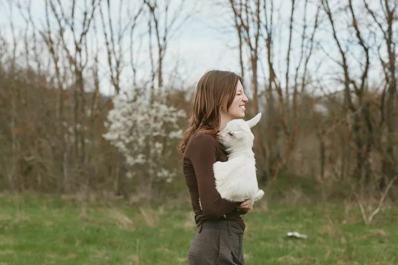 A young woman in a brown sweater happily holds a white baby goat in a green field during early spring, enjoying a peaceful moment in nature with blossoming trees in the background.