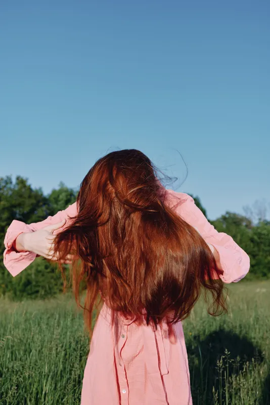 Young woman with flowing red hair in a pink dress enjoying nature against a green backdrop, representing freedom and natural beauty