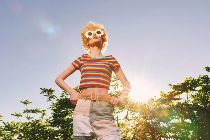 Vibrant woman wearing striped crop top and patchwork shorts, flower sunglasses, stands outdoors under bright sun, conveying carefree summer lifestyle with retro film tones.