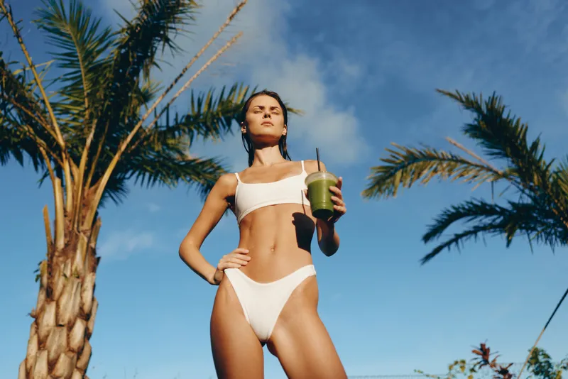 A confident woman in a white bikini stands outdoors under palm trees, holding a smoothie cup. A bright, sunny lifestyle moment emphasizing summer fitness, self care, and leisure.