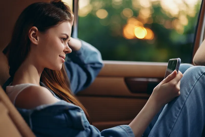 Young woman in casual denim outfit enjoying a phone call in a car during sunset, conveying feelings of happiness and connection with nature.