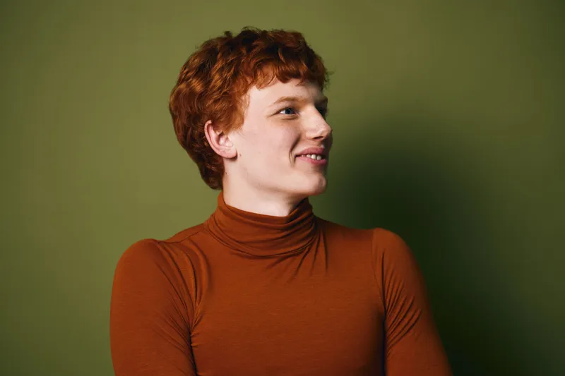 Young man with red hair wearing brown turtleneck sweater smiling and looking to side against green background in studio portrait.