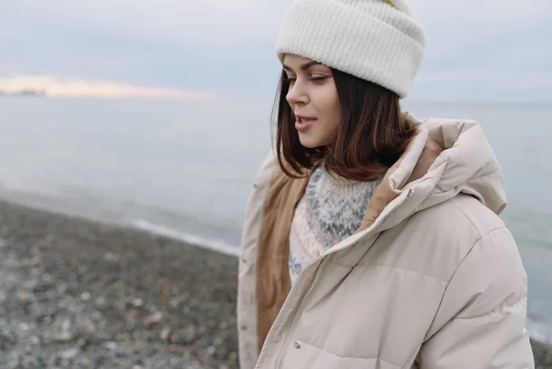 Woman in a warm coat and beanie stands by a calm beach, winter scenery and quiet mood. Soft light, chilly breeze, distant horizon, reflective moment outdoors.