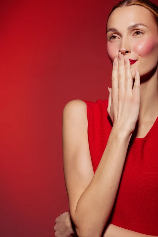 Young woman in red sleeveless top covers mouth with hand, smiling shyly against red background. Concept of emotion, beauty, fashion, and candid moment.