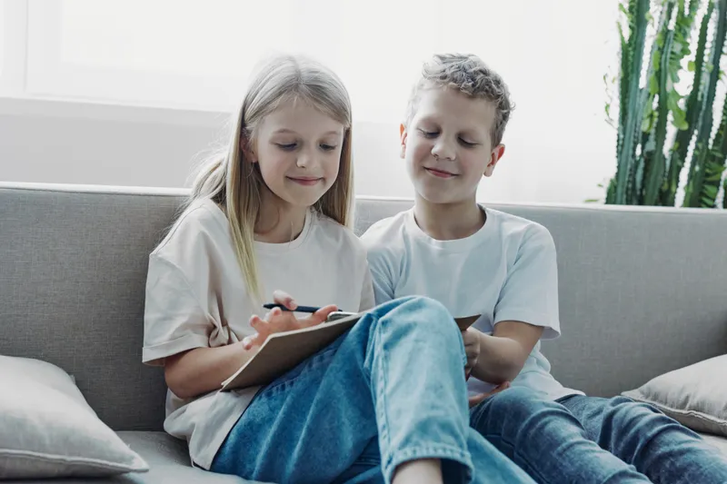 Two children sitting on a sofa, girl writing on a clipboard, boy watching attentively, casual home environment, natural light, relaxed mood.