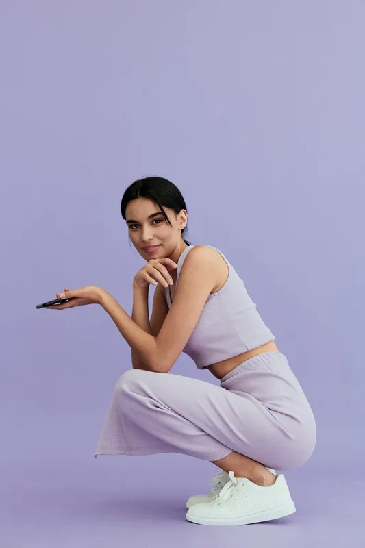 young woman wearing casual lilac outfit squatting and holding remote control against purple background, stylish pose and relaxed expression