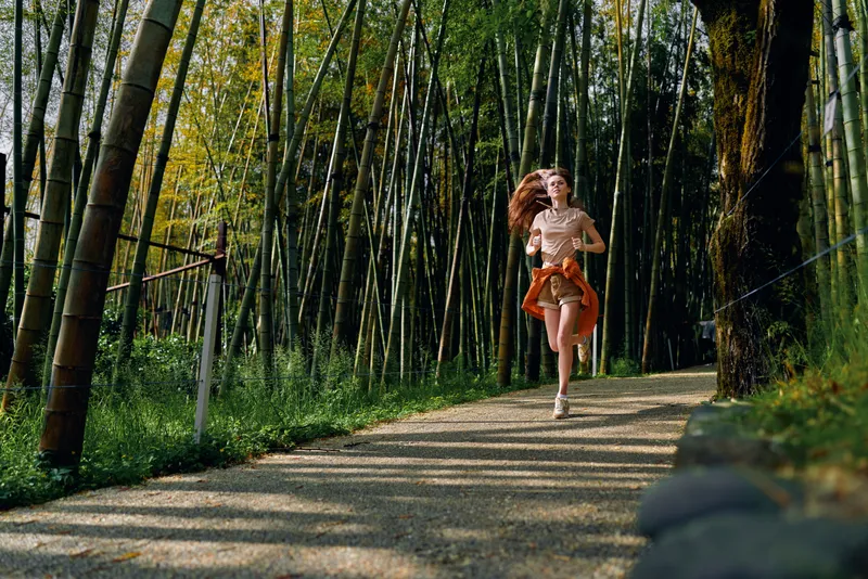 Woman running on a gravel path through a dense bamboo forest, jogging in nature on a sunny morning. Fitness motion, trail exercise, long hair and outdoor healthy lifestyle in green grove.
