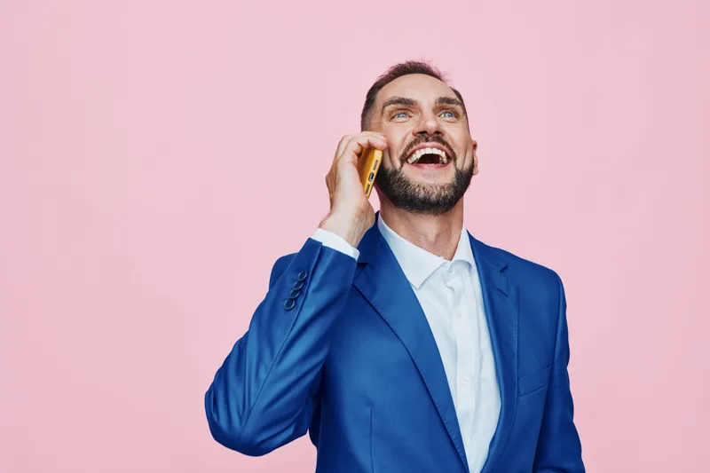 Professional business man stands against a solid pink background, isolated color, smiling as he talks on the phone. The scene conveys confidence, optimism, and approachable leadership in a modern