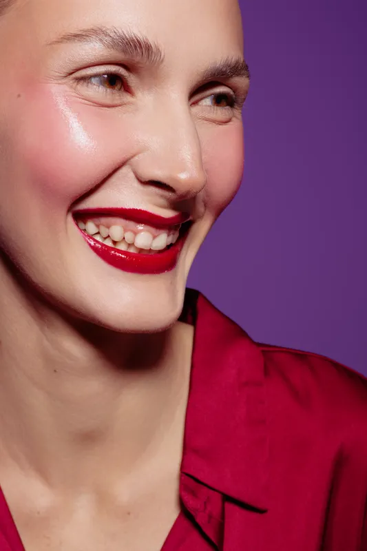 Close-up of smiling woman with red lipstick and glowing skin wearing red shirt on purple background. Concept of beauty, makeup, skincare, happiness, and fashion portrait.