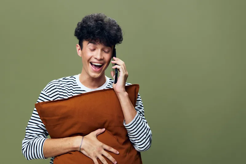 Smiling man talking on phone while holding brown pillow, wearing striped shirt, expressing joy and laughter against olive green background, casual indoor setting.