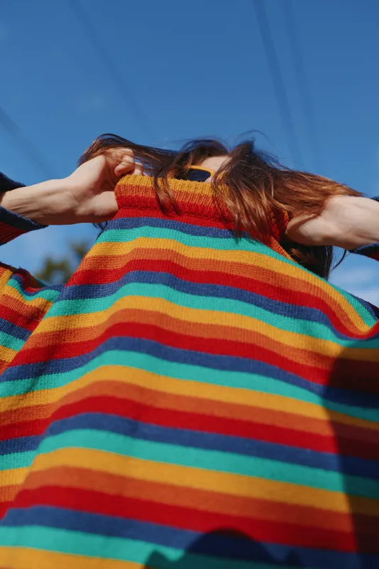Woman in her 20s pulling a colorful striped sweater over her face, casual outfit with long hair, playful expression, outdoor under clear blue sky and warm sunlight for lifestyle fashion.