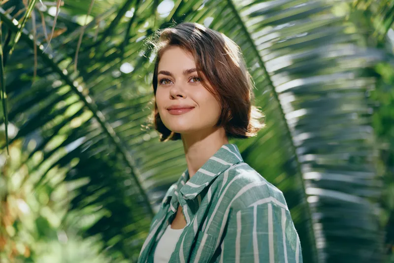 Portrait of a young woman outdoors among tropical plants, smiling softly, wearing a striped green shirt over a white top, natural sunlight.
