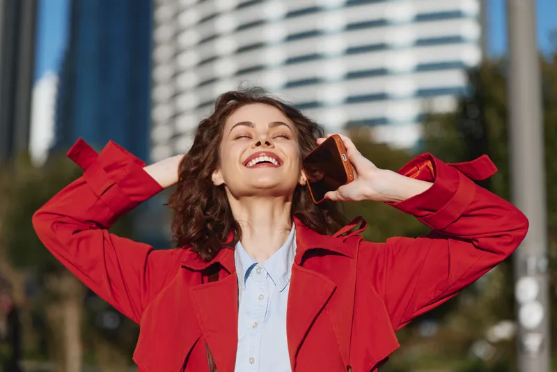 Happy woman outdoors smiling with eyes closed holding smartphone in red coat sunny day urban background casual lifestyle
