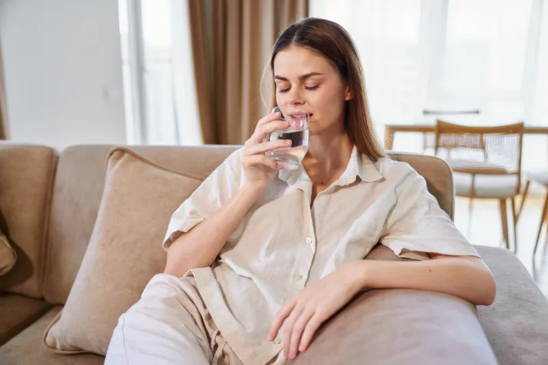 Woman drinking water on a sofa at home, relaxed and comfortable in casual clothes, portrait of hydration and wellness in a serene living room with natural light.