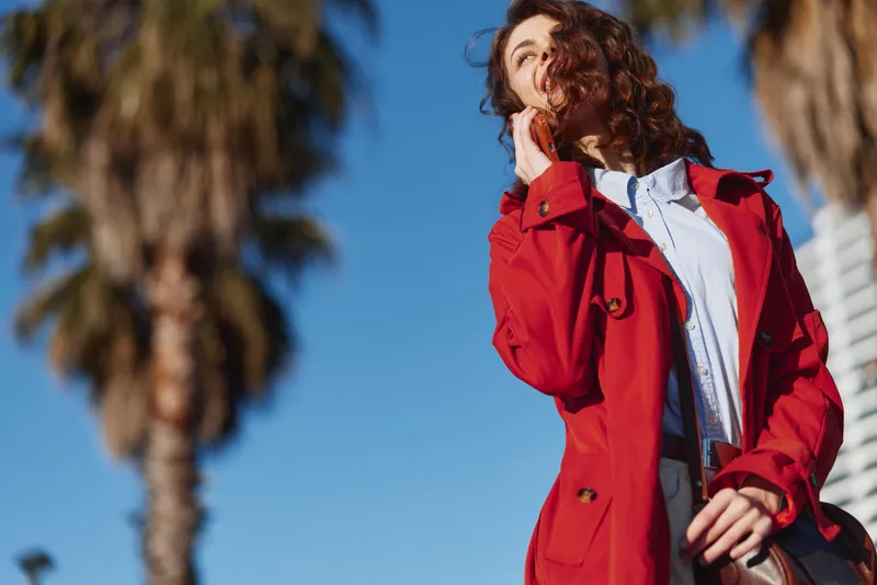 Smiling woman with curly hair wearing a red coat outdoors under bright sunlight, a fashion portrait capturing confidence, style, warmth, and carefree mood.