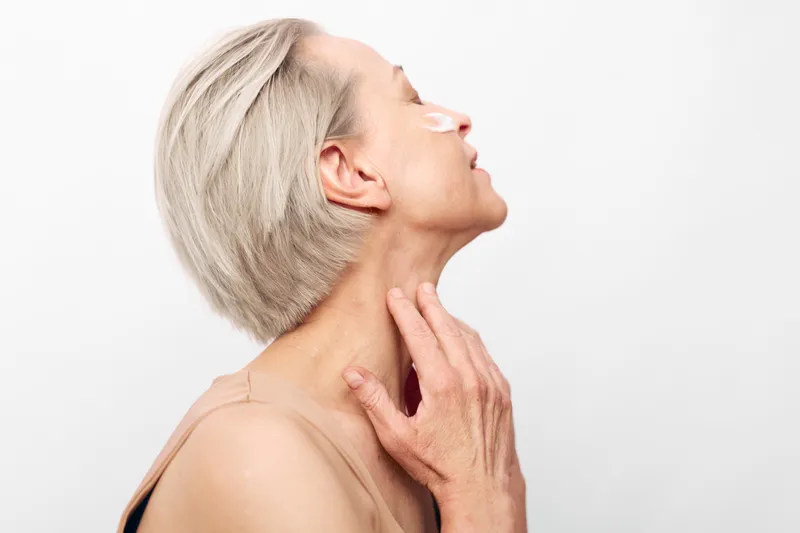 A mature woman with short grey hair applies white cream under her eyes, showing skincare routine and self-care in a bright studio with white background. Concept of aging and beauty.