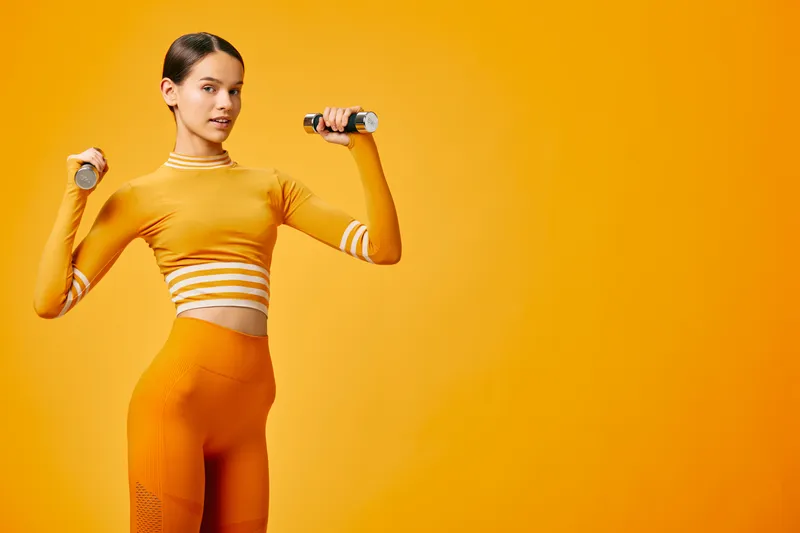 Fit young woman in orange activewear lifting small dumbbells in a studio sport pose, toned midriff visible, focused expression, minimalist monochrome orange background.