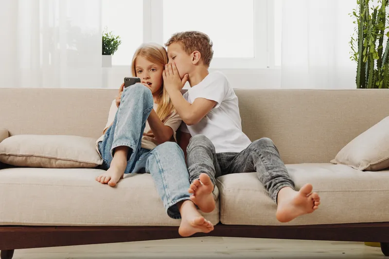 Two children sitting on a beige sofa, a boy whispering a secret to a girl who is holding a smartphone. Natural light brightens the cozy living room with plants in the background.