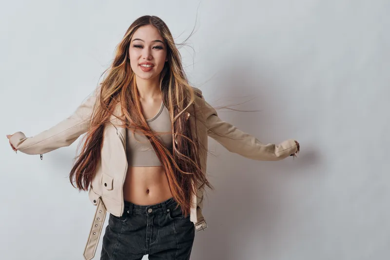 Young woman with long hair wearing casual jacket and crop top posing on gray background. Female model with closed eyes and relaxed posture in studio fashion photography.