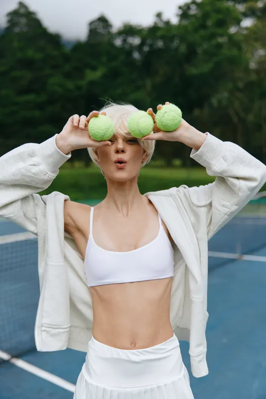 Playful young woman enjoying tennis outdoors, wearing a white crop top and skirt, smiling with tennis balls in hand, capturing vibrant summer energy