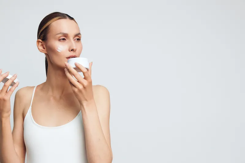 Young woman with clean skin applies moisturizing cream on her face holds jar of skincare product in hand isolated on light background with natural beauty and healthy lifestyle concept