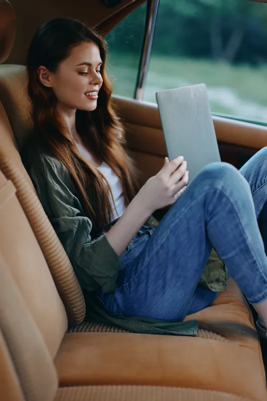 Young woman enjoying a tablet in a vintage car, dressed casually in denim jeans and a green jacket, radiating happiness in a serene outdoor setting.