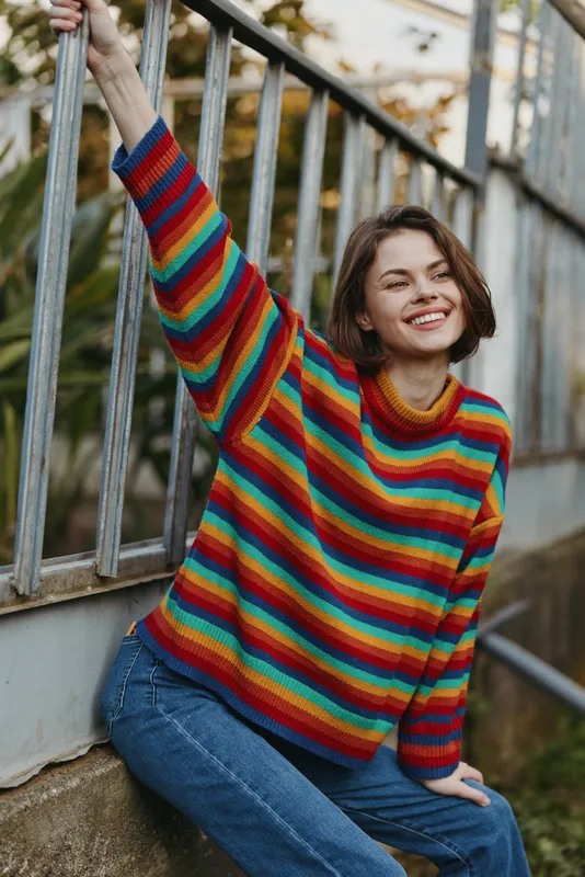 Woman smiling in a colorful rainbow sweater, casual jeans in an outdoor portrait. Young female sits by metal railing with cozy knit, relaxed pose and cheerful expression.