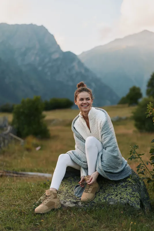 Cheerful woman outdoors, sitting on a rock in a meadow, wearing knitwear and boots, smiling at the camera, sunlit mountains in the background, relaxed lifestyle moment