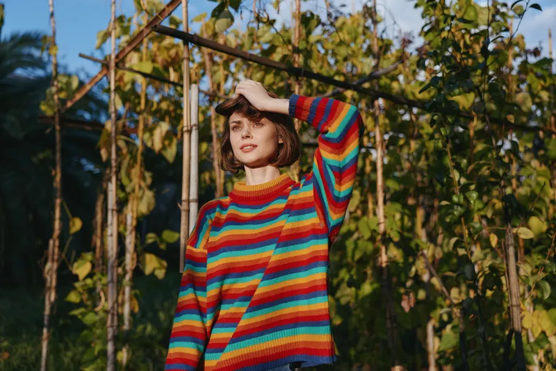 Woman in a striped sweater and casual outfit standing in a garden with sunlight, smiling, short hair, tropical outdoor vines, relaxed pose for lifestyle travel portrait.