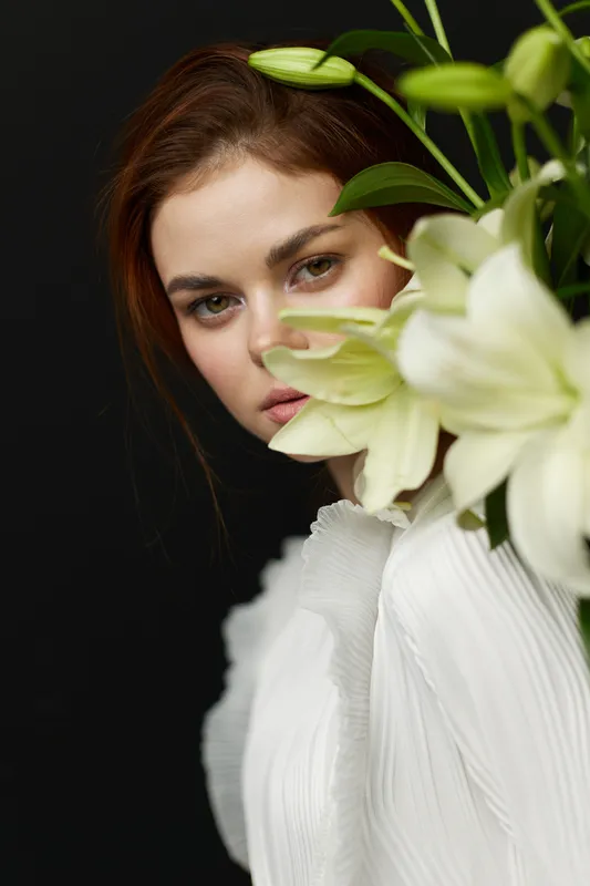 Portrait of a young Caucasian woman with long brown hair, holding white lilies against a dark background, expressing serene beauty and elegance