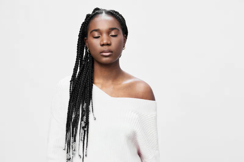 Serene young woman with braided hair wearing a cozy white sweater against a soft gray background, embodying calm and tranquility with closed eyes
