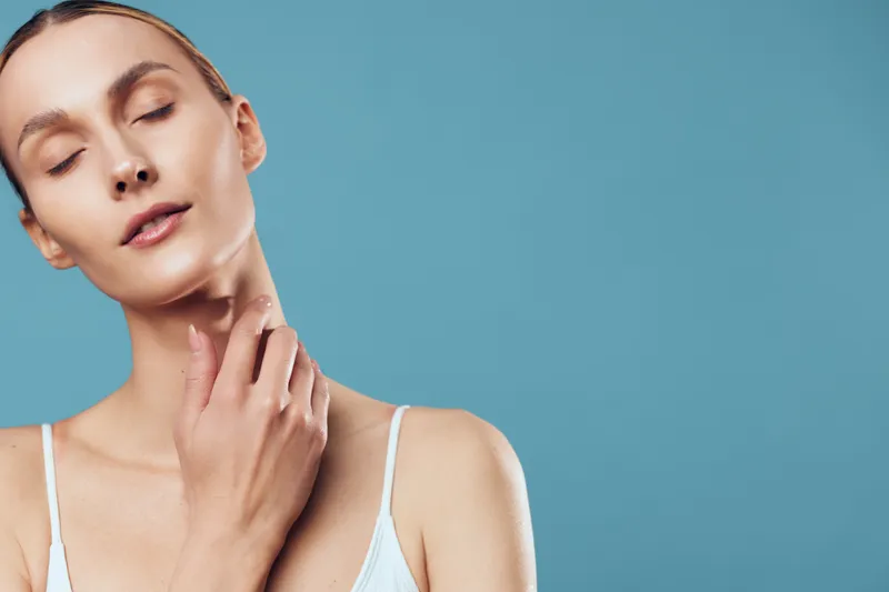 Calm woman with eyes closed touching neck, soft lighting, serene mood, blue background, warm skin tones, minimalist pose, natural expression, intimate portrait, relaxed ambiance