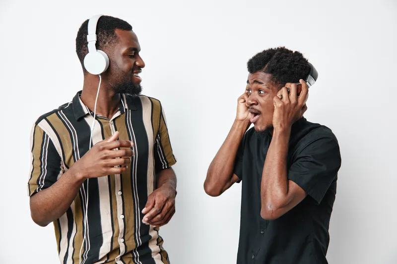Two young black men with dark skin wearing headphones enjoying music and smiling together in casual clothes on white background. Friends having fun and sharing audio experience.
