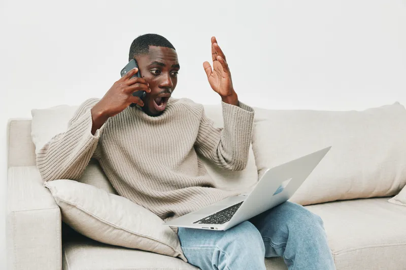 Young Black man expressing surprise while on the phone and using a laptop at home with a neutral background, showcasing emotions of excitement and engagement