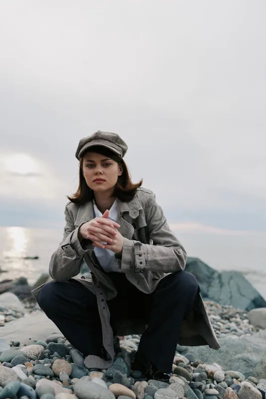 Young woman in a checkered coat and cap squats on a rocky beach looking thoughtful under an overcast sky. Concept of fashion, autumn, and outdoor lifestyle.